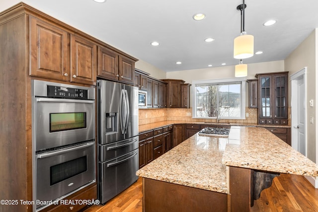 kitchen with light stone counters, backsplash, appliances with stainless steel finishes, light wood finished floors, and glass insert cabinets
