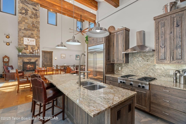 kitchen featuring a fireplace, a towering ceiling, wall chimney range hood, appliances with stainless steel finishes, and tasteful backsplash