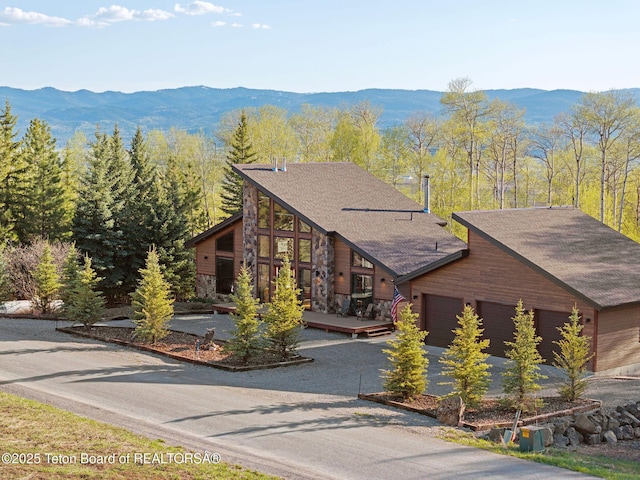 log-style house featuring an attached garage, stone siding, a shingled roof, driveway, and a deck with mountain view