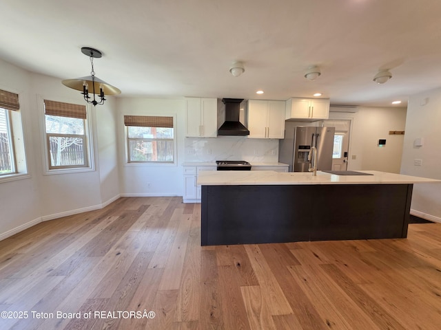 kitchen with wall chimney range hood, a kitchen island with sink, stainless steel refrigerator with ice dispenser, white cabinetry, and light countertops