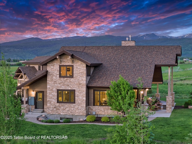 1795 Fumarole Dr, Victor, ID 83455 - view of front of property featuring stone siding, a mountain view, a chimney, roof with shingles, and a front lawn