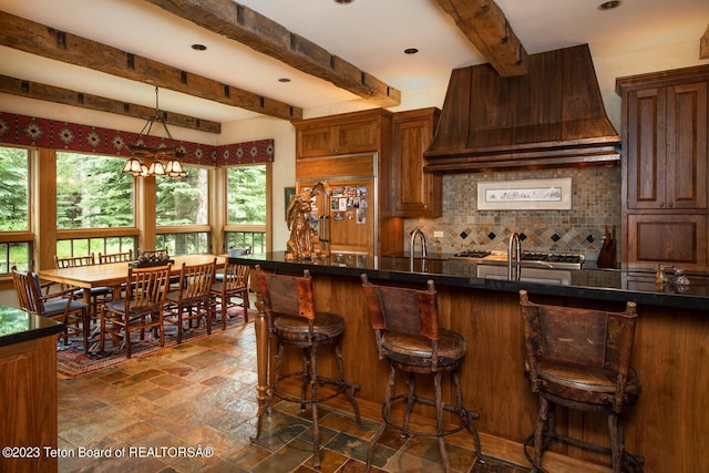 kitchen with decorative backsplash, beamed ceiling, premium range hood, dark countertops, and a kitchen bar