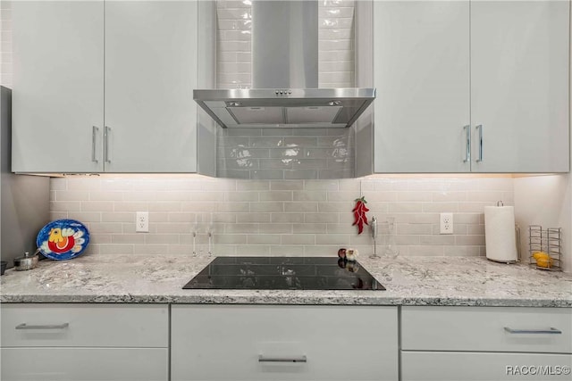 kitchen with wall chimney range hood, backsplash, black electric stovetop, light stone countertops, and white cabinets