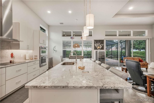 kitchen featuring sink, wall chimney range hood, hanging light fixtures, black electric stovetop, and a kitchen bar