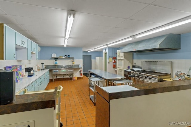 kitchen with blue cabinets, oven, light tile patterned floors, kitchen peninsula, and wall chimney exhaust hood