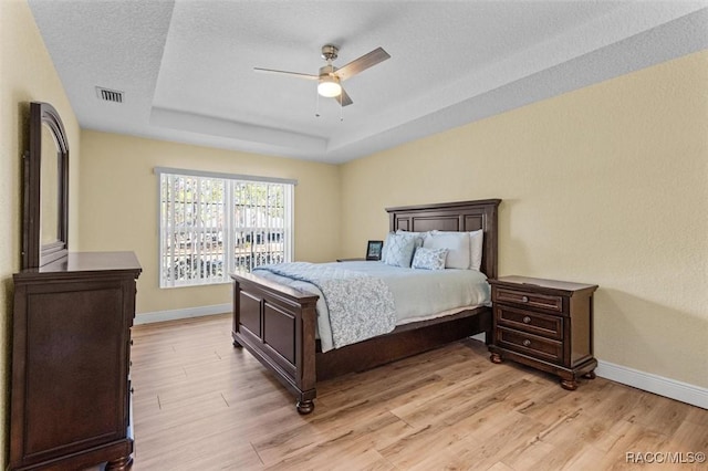 bedroom with light wood-type flooring, a tray ceiling, a textured ceiling, and ceiling fan