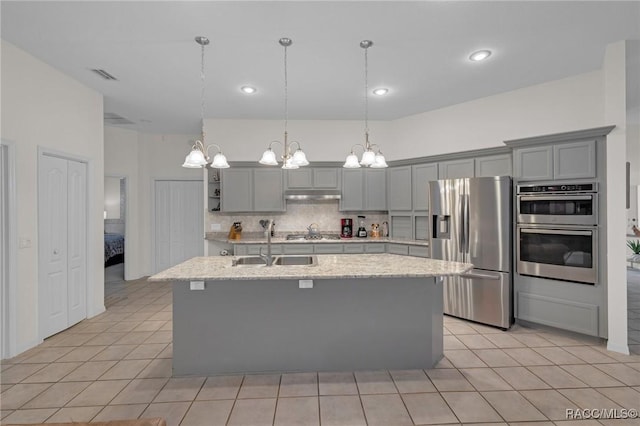 kitchen with gray cabinetry, under cabinet range hood, stainless steel appliances, an island with sink, and decorative light fixtures