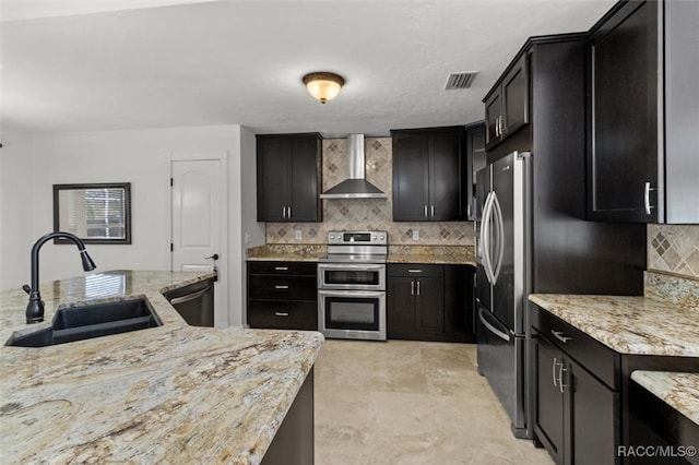 kitchen featuring decorative backsplash, wall chimney exhaust hood, light stone countertops, stainless steel appliances, and a sink