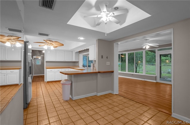 kitchen with white cabinetry, stainless steel refrigerator with ice dispenser, kitchen peninsula, light hardwood / wood-style floors, and a tray ceiling