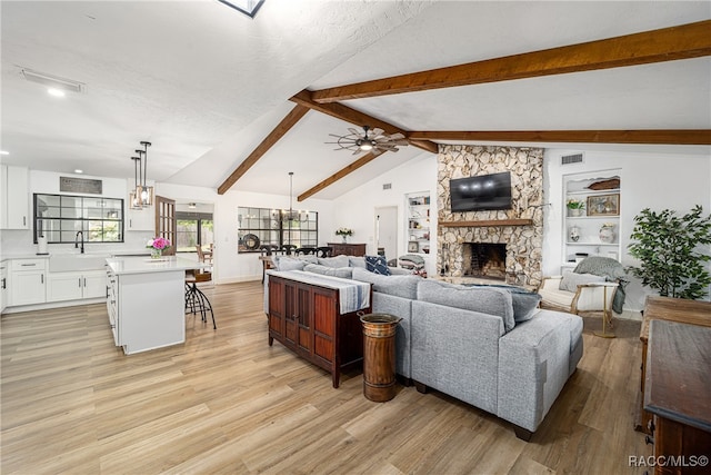 living room with ceiling fan with notable chandelier, sink, lofted ceiling with beams, light hardwood / wood-style floors, and a stone fireplace