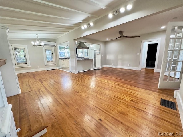 unfurnished living room featuring ceiling fan with notable chandelier, beamed ceiling, light hardwood / wood-style floors, and a wall mounted air conditioner