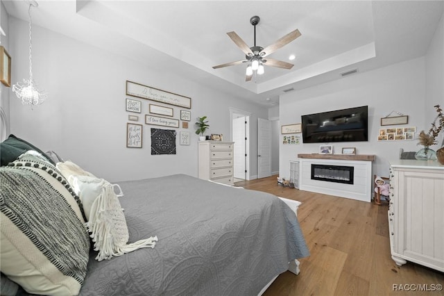 bedroom featuring ceiling fan with notable chandelier, wood finished floors, visible vents, a tray ceiling, and a glass covered fireplace