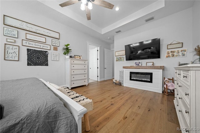 bedroom featuring ceiling fan, hardwood / wood-style flooring, visible vents, a tray ceiling, and a glass covered fireplace