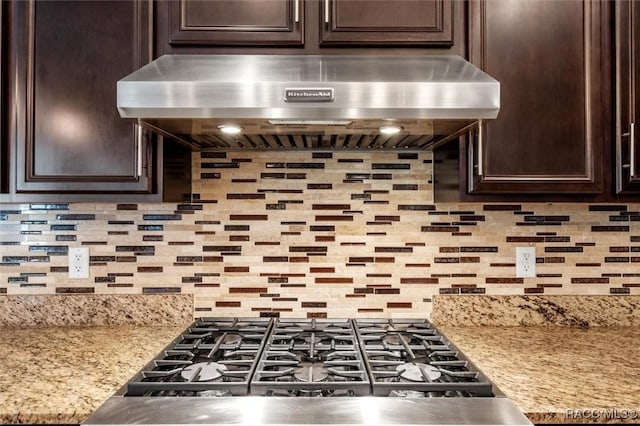 kitchen with light stone countertops, backsplash, dark brown cabinetry, and range hood