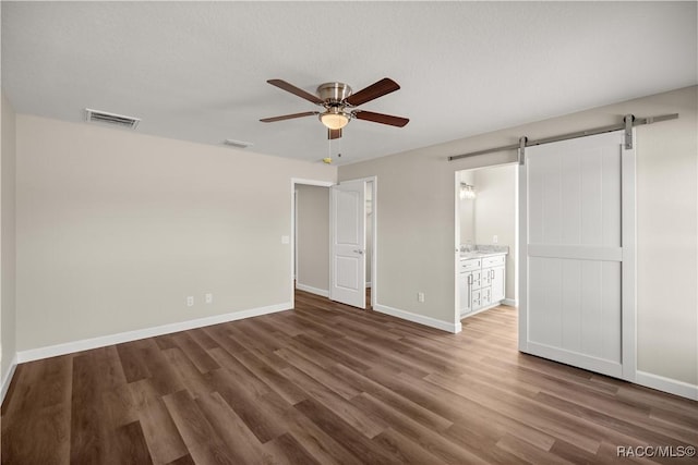 unfurnished bedroom featuring dark hardwood / wood-style flooring, ensuite bath, a barn door, and ceiling fan