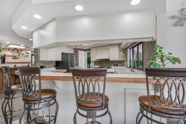 kitchen with fridge with ice dispenser, white cabinets, a sink, custom range hood, and backsplash