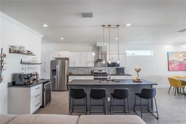 kitchen with appliances with stainless steel finishes, white cabinetry, a kitchen island with sink, beverage cooler, and wall chimney exhaust hood