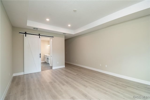 unfurnished bedroom featuring a raised ceiling, a barn door, ensuite bath, and light hardwood / wood-style flooring