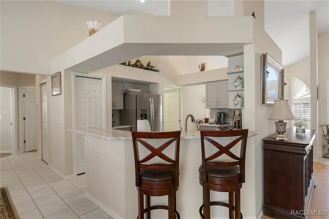 kitchen with a peninsula, light tile patterned floors, gray cabinetry, stainless steel refrigerator with ice dispenser, and a kitchen bar