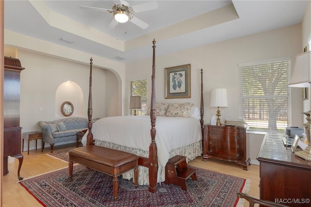 bedroom featuring a tray ceiling, multiple windows, arched walkways, and wood finished floors