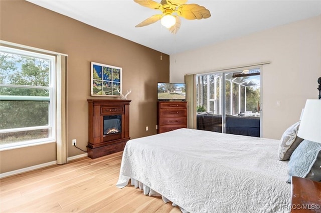 bedroom featuring access to exterior, light wood-style flooring, a ceiling fan, a glass covered fireplace, and baseboards