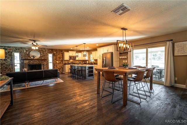 dining area with ceiling fan with notable chandelier, a textured ceiling, dark wood-type flooring, sink, and a stone fireplace