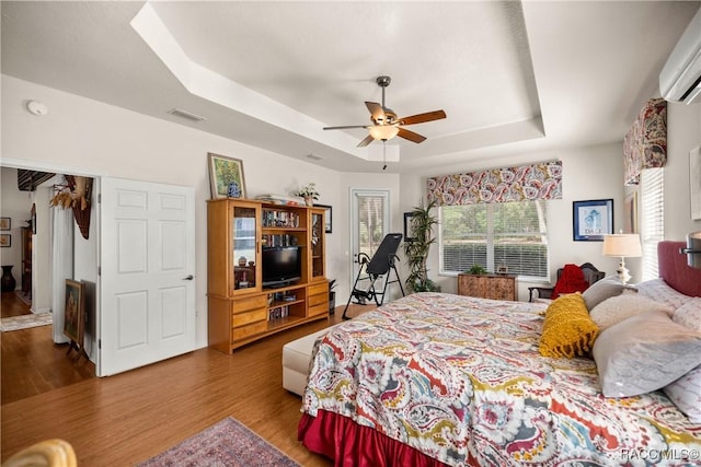 bedroom with hardwood / wood-style floors, a wall mounted AC, ceiling fan, and a tray ceiling