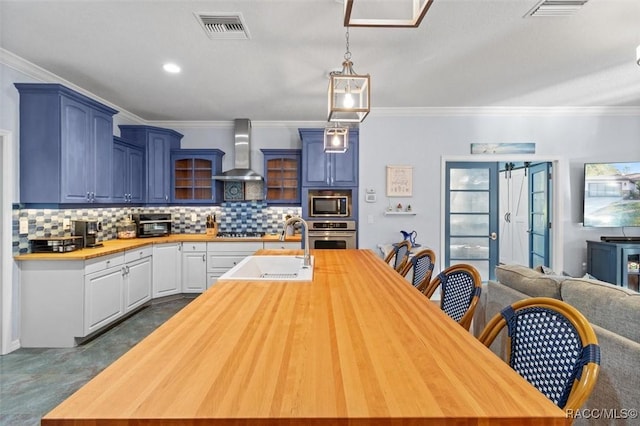 kitchen with appliances with stainless steel finishes, wall chimney exhaust hood, blue cabinets, sink, and hanging light fixtures