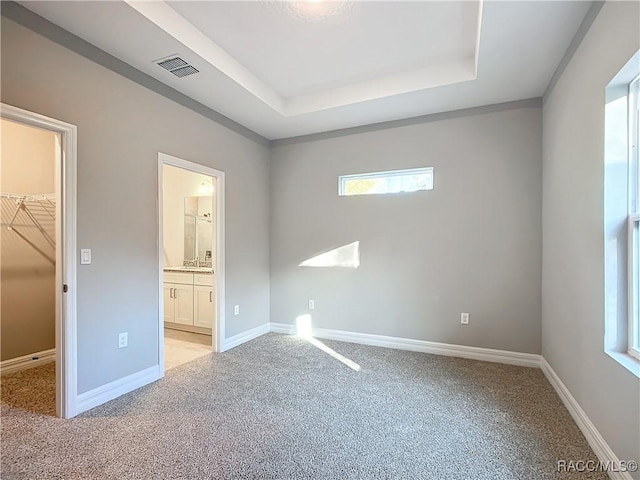 unfurnished bedroom with ensuite bathroom, a spacious closet, a raised ceiling, a closet, and light colored carpet