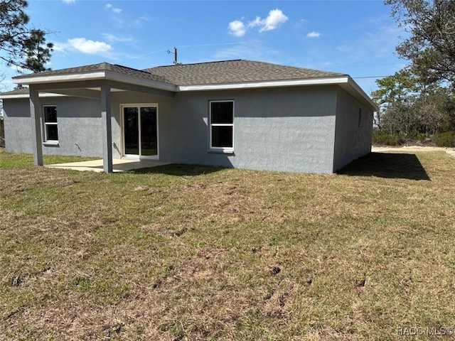 rear view of house with stucco siding, roof with shingles, a lawn, and a patio