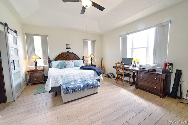 bedroom with light wood-type flooring, a barn door, a raised ceiling, and a ceiling fan