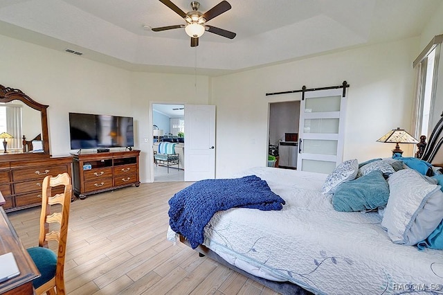 bedroom with a barn door, visible vents, a raised ceiling, ceiling fan, and light wood-style floors