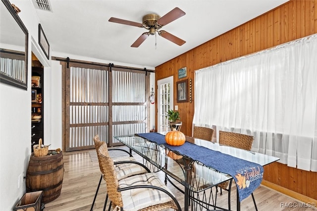 dining room with a barn door, ceiling fan, light hardwood / wood-style floors, and wood walls