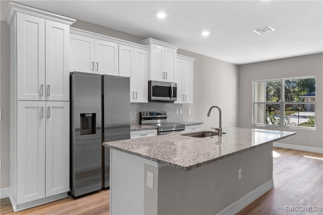 kitchen with light stone counters, a center island with sink, stainless steel appliances, light wood-style flooring, and a sink