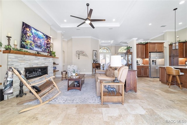 living room featuring ceiling fan, ornamental molding, a tray ceiling, and a stone fireplace