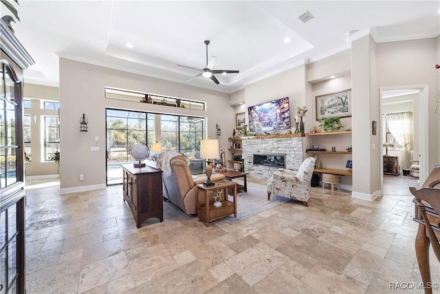 living room with a stone fireplace, ornamental molding, ceiling fan, a raised ceiling, and built in shelves