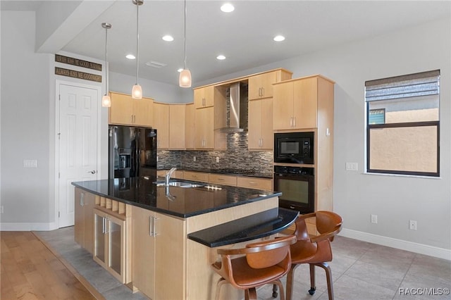 kitchen featuring light brown cabinetry, decorative light fixtures, an island with sink, black appliances, and wall chimney exhaust hood