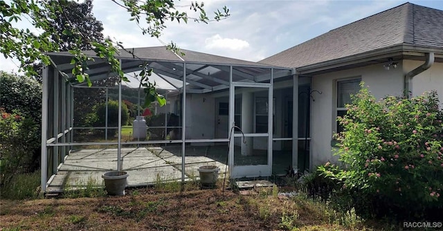 rear view of property featuring stucco siding, a shingled roof, glass enclosure, and a patio