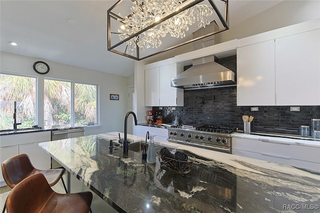 kitchen with stainless steel appliances, sink, white cabinetry, dark stone counters, and wall chimney range hood