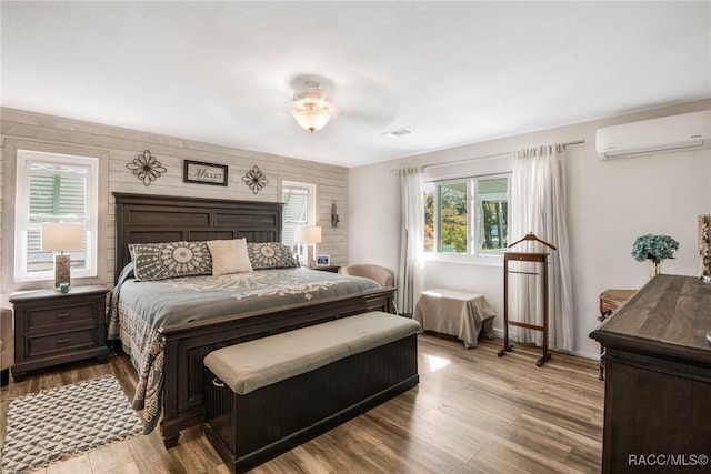 bedroom featuring a wall mounted air conditioner, ceiling fan, wood walls, and light wood-type flooring