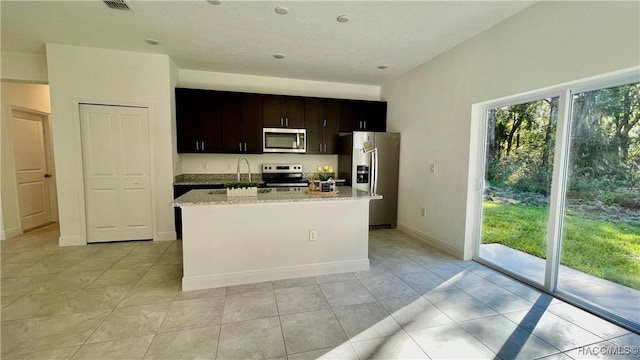 kitchen with light stone counters, a kitchen island with sink, dark brown cabinetry, stainless steel appliances, and baseboards