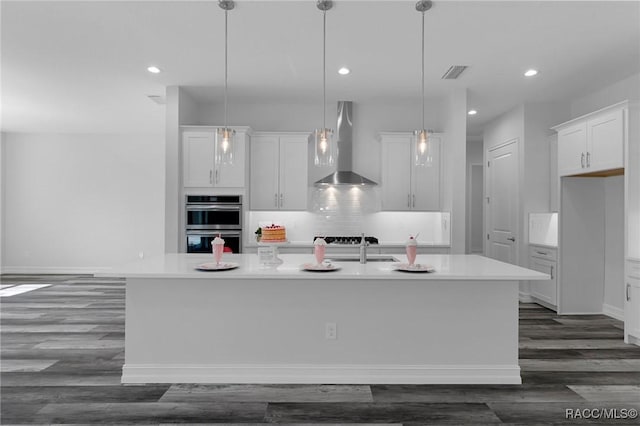 kitchen featuring a center island with sink, backsplash, double oven, white cabinets, and wall chimney exhaust hood