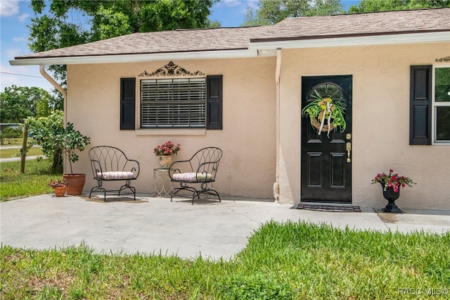 view of exterior entry featuring a shingled roof, stucco siding, and a patio