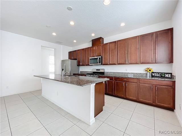 kitchen featuring sink, an island with sink, dark stone counters, light tile patterned floors, and appliances with stainless steel finishes