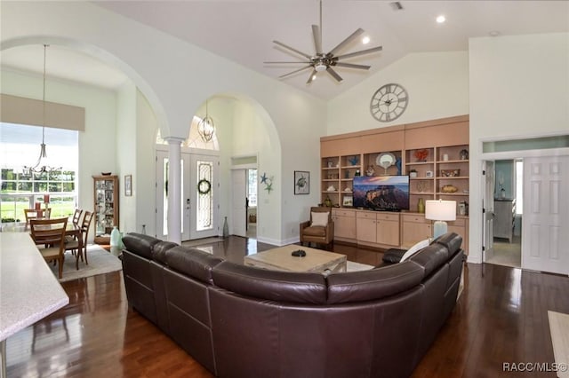 living room with ornate columns, ceiling fan with notable chandelier, high vaulted ceiling, dark hardwood / wood-style flooring, and built in shelves