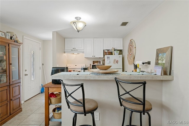 kitchen with light tile patterned floors, under cabinet range hood, white appliances, white cabinets, and a kitchen bar