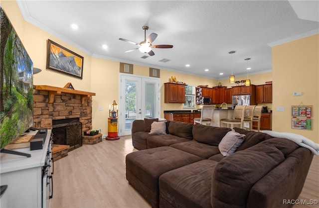 living room featuring french doors, light wood-type flooring, ornamental molding, ceiling fan, and a stone fireplace