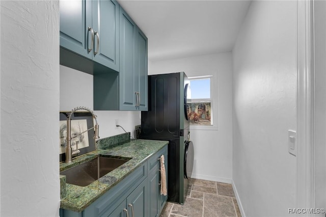 kitchen with stacked washing maching and dryer, blue cabinetry, dark stone counters, and sink