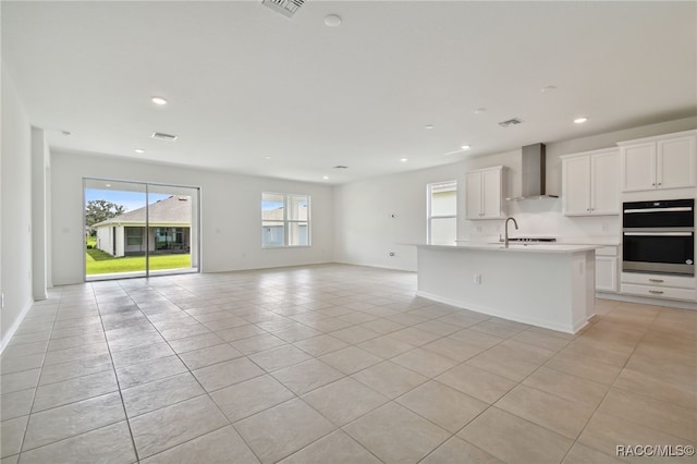 kitchen with wall chimney range hood, light tile patterned flooring, double oven, a center island with sink, and white cabinets