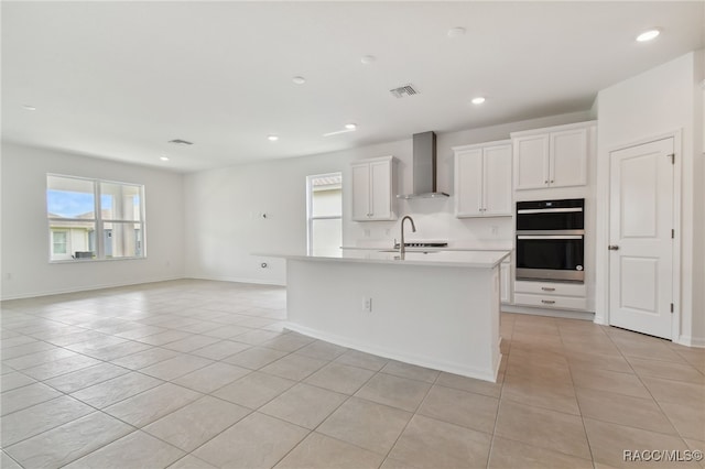 kitchen with wall chimney exhaust hood, stainless steel double oven, an island with sink, white cabinetry, and light tile patterned flooring